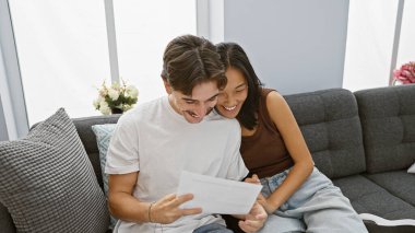 Mutlu bir ırklar arası çift modern bir oturma odasındaki kanepede birlikte doküman okuyor, neşe ve beraberliği ifade ediyor..