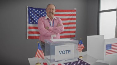 Orta yaşlı ve bıyıklı bir adam, Amerikan bayrağı ve oy verme kabininin önünde kollarını kavuşturup, güvenle kapalı bir yerde duruyor..