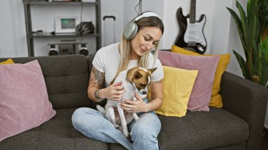 Beyaz kadın kulaklıkla müzik dinlerken Jack Russell 'ın köpeğini rahat bir kanepede kucağında tutuyor..