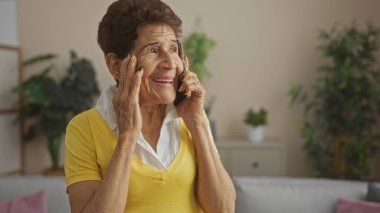 Kısa saçlı, evde konuşan yaşlı İspanyol kadın. Bitkilerle dolu bir salonda oturmuş gülümsüyor..