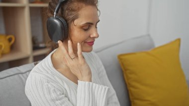 İçerik sahibi genç bir kadın, rahat bir oturma odası ortamında kulaklıkla müzik dinlemekten zevk alır. Rahatlık ve rahatlık yayar..