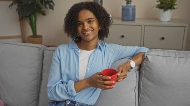 Güzel, Afro-Amerikan bir kadın, kıvırcık saçlı, sıcak bir oturma odasında elinde kırmızı bardakla oturuyor, mavi bir gömlek ve beyaz bir saat giyiyor, rahat bir şekilde gülümsüyor..