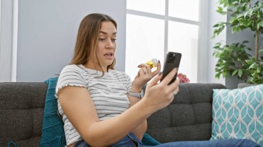 Genç bir kadın modern bir oturma odasında telefonuna danışarak ilaçlarını inceliyor, sağlık ve teknoloji entegrasyonunu gösteriyor..