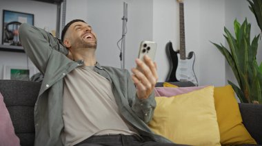 Rahat İspanyol adam akıllı telefon kullanıyor ve rahat bir oturma odasında gülüyor..