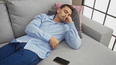 Sakallı yakışıklı, genç, İspanyol bir adam evinin oturma odasındaki kanepede uyuyor. Yanında da akıllı telefonu var..