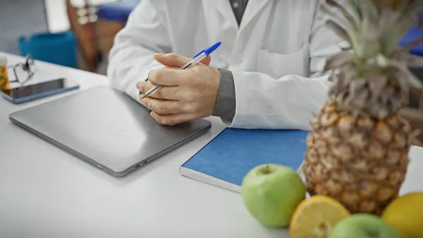 stock image Close-up of a man in a white coat writing on a clipboard in a health clinic, with healthy fruits in the background.