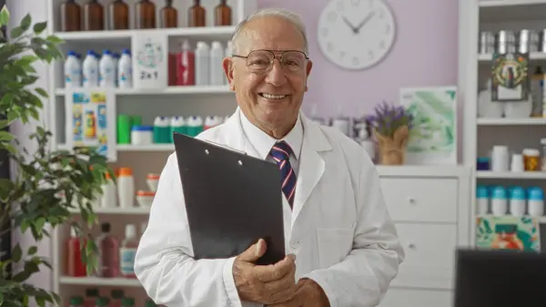 stock image Elderly grey-haired man smiles while holding a clipboard in a pharmacy room filled with colorful products and shelves in the background.