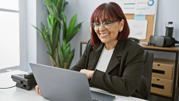 stock image A smiling mature hispanic woman working on a laptop in a modern office setting.