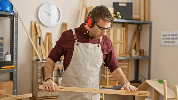 A focused man with a beard wearing safety earmuffs and an apron measures wood in a well-organized carpentry workshop.
