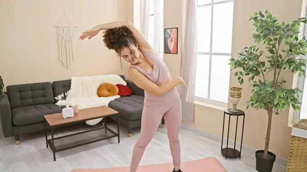 stock image A young woman with curly hair exercises in her modern living room, stretching on a pink yoga mat.