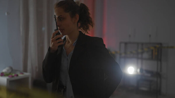 Молодая белая женщина расследует тускло освещенное место преступления, общаясь по радио, излучая чувство срочности и сосредоточенности..