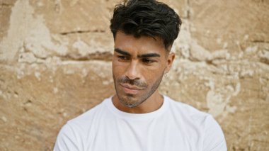 Yakışıklı, sakallı, beyaz tişörtlü, kahverengi tuğlalı bir arka planı olan bir İspanyol..