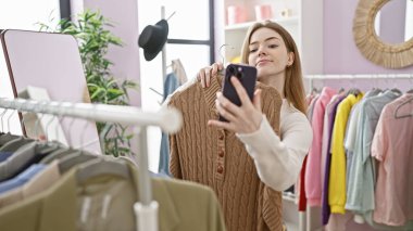 Sarışın kadın renkli bir elbise odasında, akıllı telefonuyla selfie çekiyor. Yaşam tarzını ve modasını yansıtıyor..