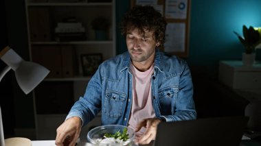 Ofiste çalışırken yemek yiyen İspanyol bir adam, dizüstü bilgisayar, günlük kıyafetler, loş bir iç mekan..