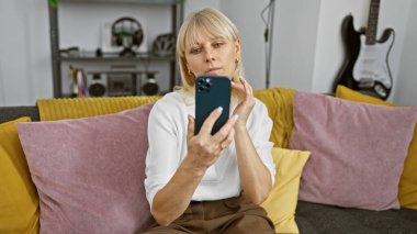 Modern oturma odasında arka planda gitarıyla akıllı telefon kullanan beyaz bir kadın.