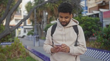 Afrika kökenli Amerikalı bir adam, ağaçlı ve fayanslı bir bankta yürürken akıllı telefon kullanıyor..