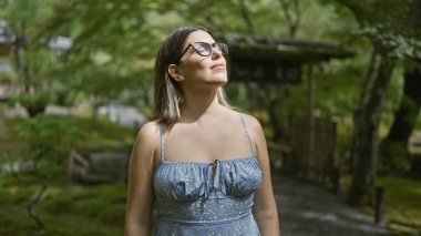 Güzel İspanyol kadın, gözleri kapalı, nefes nefese, Japon yeşil parkında duruyor, güneş ışığının tadını çıkarıyor, gözlük takarak meditasyon yapıyor.