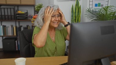 Ofiste oturan, gözlüklü yaşlı bir kadın, şaşırmış bir ifadeyle, iç mekan bitkileri ve kitaplıklarla çevrili, başını tutarken bilgisayar ekranına odaklanmış..