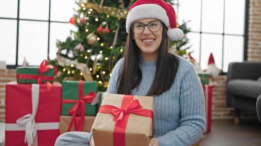 Noel şapkası takmış gülümseyen genç İspanyol kadın evde hediyesini tutuyor.