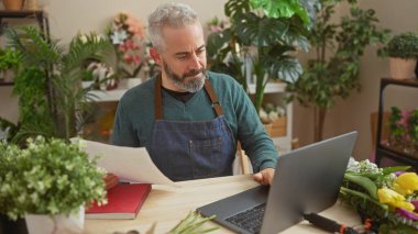 Sakallı adam yeşil bitkiler ve renkli çiçeklerle çevrili bir çiçekçide dizüstü bilgisayar kullanıyor..