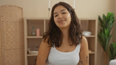 Esmer saçlı, güzel, genç bir İspanyol kadın huzur dolu bir spa odasında otururken gülümsüyor. Sağlık dekorasyonu ve iç mekan bitkileriyle..