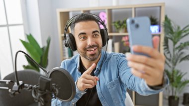 Yakışıklı bir İspanyol, modern bir radyo stüdyosunda kulaklık ve kot gömlek giyerek podcast yaparken selfie çekiyor..