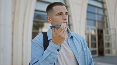 A young hispanic man in casual attire speaks on a mobile phone in front of an urban campus building clipart