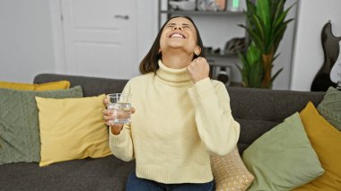 Elinde bir bardak su tutan neşeli bir kadın, renkli yastıkları olan rahat bir oturma odasında oturuyor..