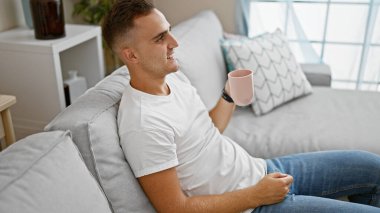 Gülümseyen genç İspanyol adam elinde kupa, rahat bir oturma odasında dinleniyor.