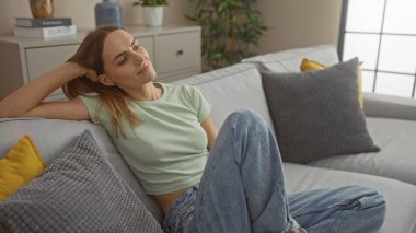 Güzel, genç, sarışın bir kadın şık bir oturma odasında rahat bir kanepede, dekoratif yastıklar ve ev bitkileriyle çevrili, evinde huzurlu bir anın tadını çıkarıyor..