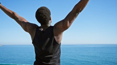 Kolları havada, güneşli sahilde özgürlüğünü kucaklayan genç, yetişkin bir siyah adamın arka görüntüsü..