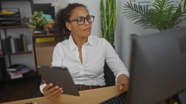 Ofiste çalışan ve elinde tablet tutan, kapalı alan bitkileri ve ofis malzemeleriyle çevrili yetişkin bir kadın, iş yerinde odaklı bir profesyoneli temsil ediyor..