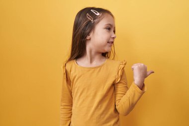 Tapılası İspanyol kız güler arkadaş canlısı, kendine güvenen, ağzı açık bir gülümseme; ayakta durur, başparmağını yalıtılmış sarı arka plan üzerinde gösterir, neşeli, pozitif, iyimser bir fikir işaret eder!