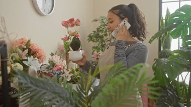 Genç bir kadın çiçekçide telefonla konuşurken bir bitkiyi inceliyor.