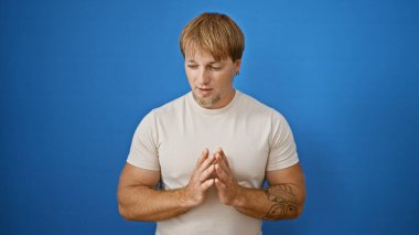 Sakallı ve dövmeli, düşünceli, genç, beyaz bir adam mavi arka planda duruyor..