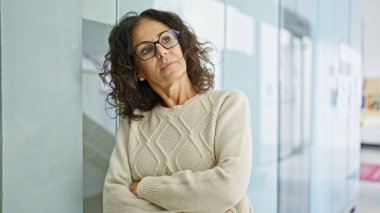 A confident middle-aged hispanic woman with glasses stands arms crossed in an office interior, exuding professionalism and experience. clipart