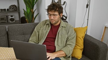 Samimi bir oturma odasında dizüstü bilgisayarda çalışırken kablosuz kulaklık kullanan sakallı İspanyol bir adam..