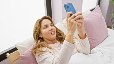 Gülümseyen İspanyol kadın yatak odasında akıllı telefon kullanıyor, evde rahatlık ve bağlılık tasvir ediyor..