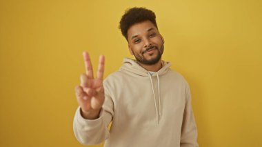 Yakışıklı siyah adam sarı arka planda bej kapüşonlu barış işareti yapıyor.