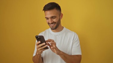 Akıllı telefon kullanırken gülümseyen İspanyol genç adam, sarı bir arka planda izole edilmiş..