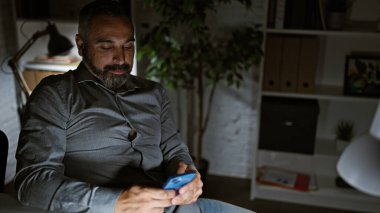 Akşamları ofiste akıllı telefon kullanan, sakallı, gri saçlı, olgun bir İspanyol..