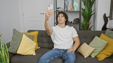 Kıvırcık saçlı İspanyol bir adam şık bir oturma odasında akıllı telefondan selfie çekiyor..