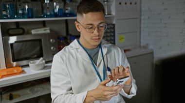 Laboratuvarda çalışan gözlüklü, laboratuvar önlüğü giyen ve akıllı telefon kullanan İspanyol bir adam.