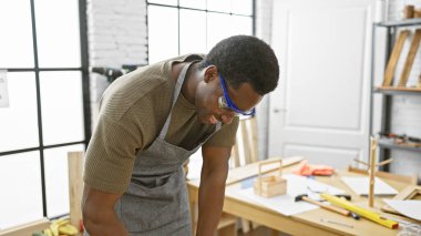 Parlak bir marangozluk atölyesinde güvenlik gözlüğü takan odaklanmış Afrikalı Amerikalı bir adam..