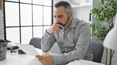 Sakallı ve gri saçlı olgun bir İspanyol modern ofis ortamında bir belgeyi incelikle gözden geçirir..