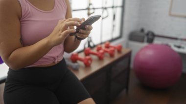 Spor salonunda telefonunu kırmızı halterli ve arka planda pembe egzersiz topuyla kontrol eden genç bir İspanyol kadın, kapalı alanda egzersiz ayarlarını vurguluyor..