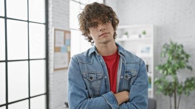 Kendine güvenen kıvırcık saçlı ve gözlüklü genç adam modern ofis ortamında kollarını kavuşturdu..