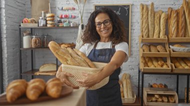 Fırında bir sepet ekmek tutan gülümseyen bir kadın raflarda çeşitli fırınlanmış mallar ve pastalarla çevrili.