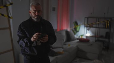 Sakallı bir adam, kapalı bir suç mahallinde cep telefonunu dikkatle inceliyor..