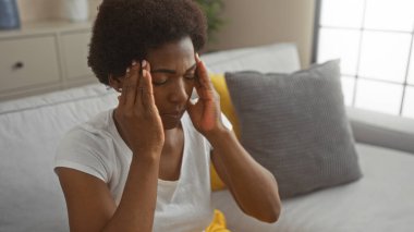 Başı ağrıyan, gözleri kapalı bir kadın, modern bir oturma odasında beyaz bir koltukta oturuyor, iki eliyle başını tutuyor..